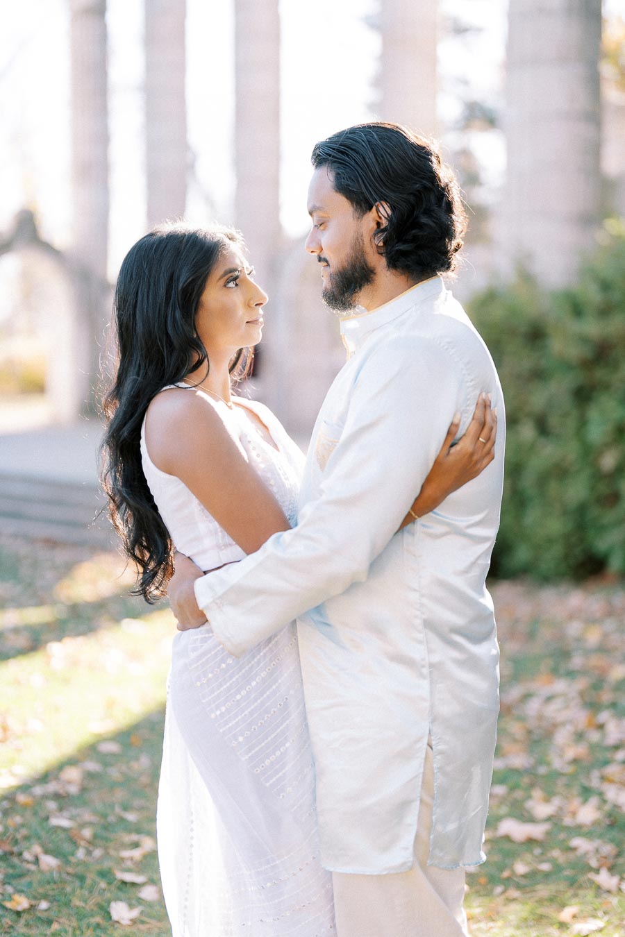 A couple in traditional white attire lovingly embraces outdoors surrounded by sunlight and greenery.