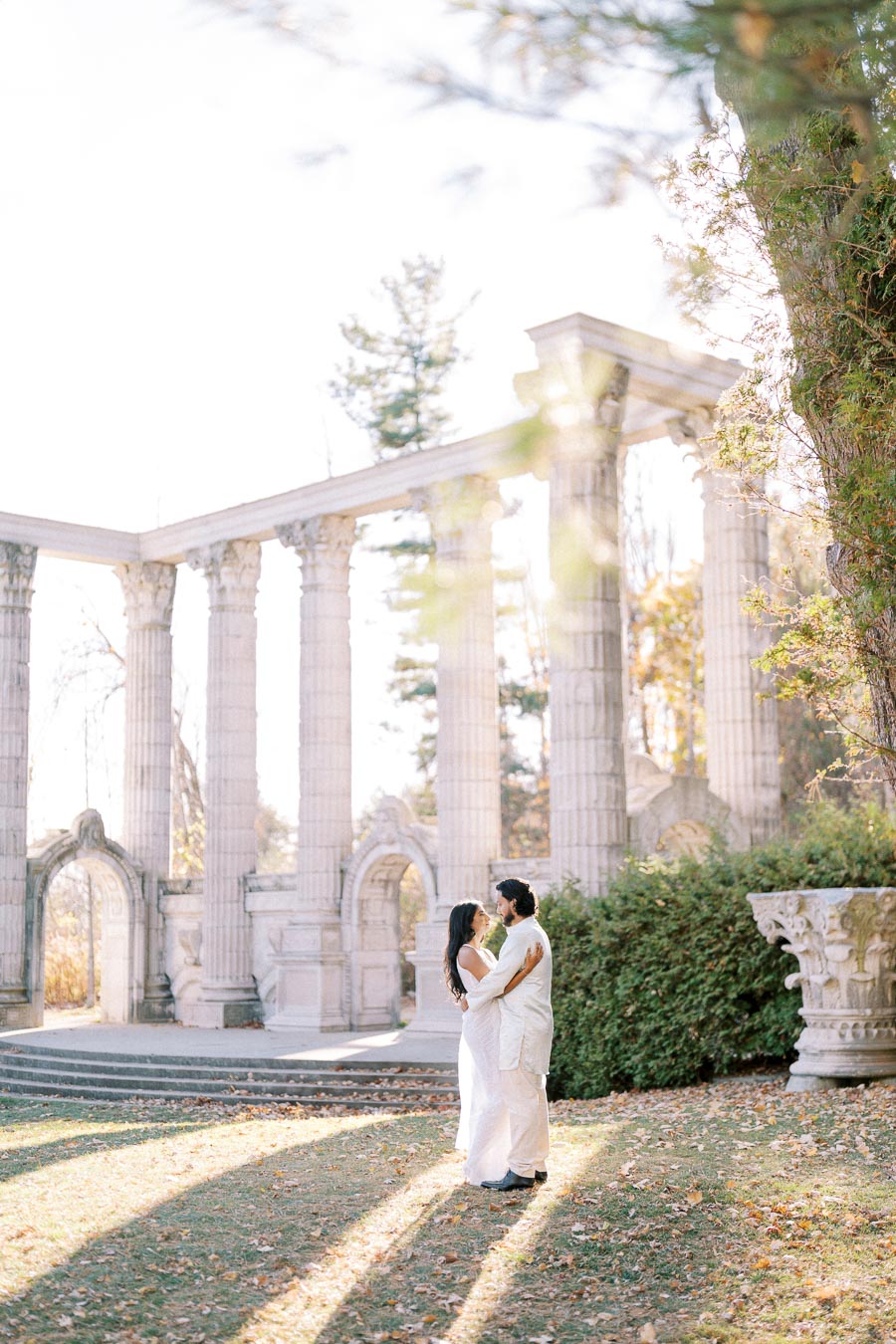 Romantic couple embracing in front of ancient architectural columns in a sunny garden setting.