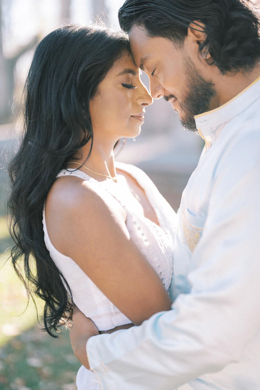 A couple embracing tenderly outdoors, eyes closed, with soft sunlight enhancing the romantic atmosphere. The woman has long dark hair and the man a short beard. Both wear light-colored clothing.