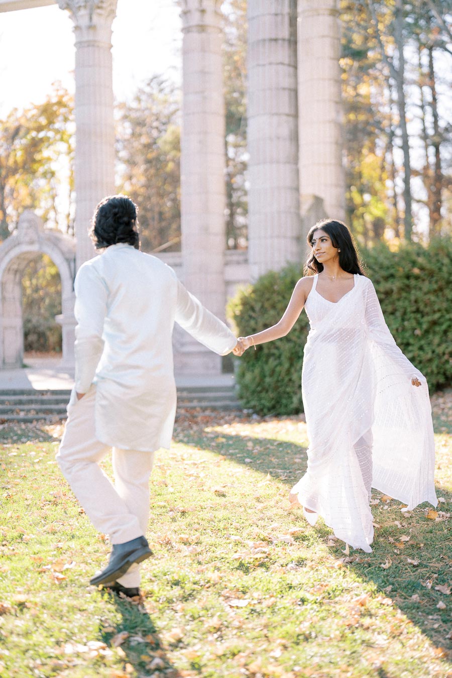 Couple in white attire holding hands in a sunlit garden setting with stone columns, surrounded by autumn foliage.