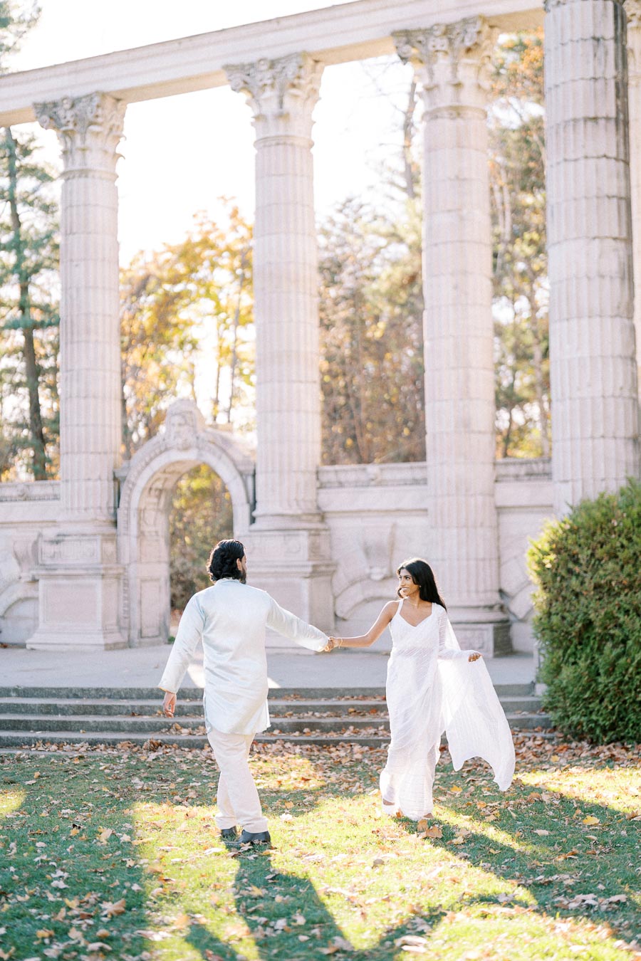 A couple in white traditional attire joyfully holding hands and walking on grass with a backdrop of majestic white columns and autumn trees.