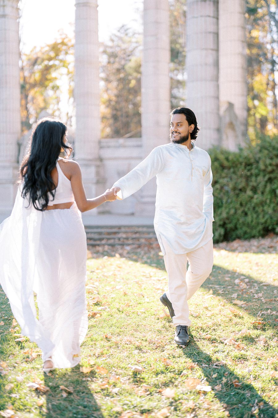 A couple in elegant white attire enjoying a romantic moment in a sunlit garden with towering stone pillars in the background, embracing the warmth of a beautiful autumn day.