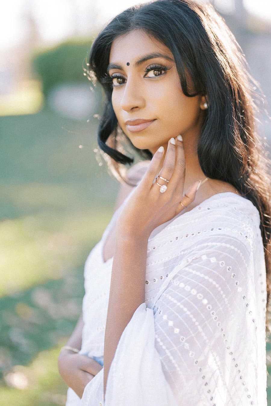 Young woman in a white sari with intricate patterns, standing outdoors with soft sunlight illuminating her face, showcasing a serene expression and elegant jewelry.