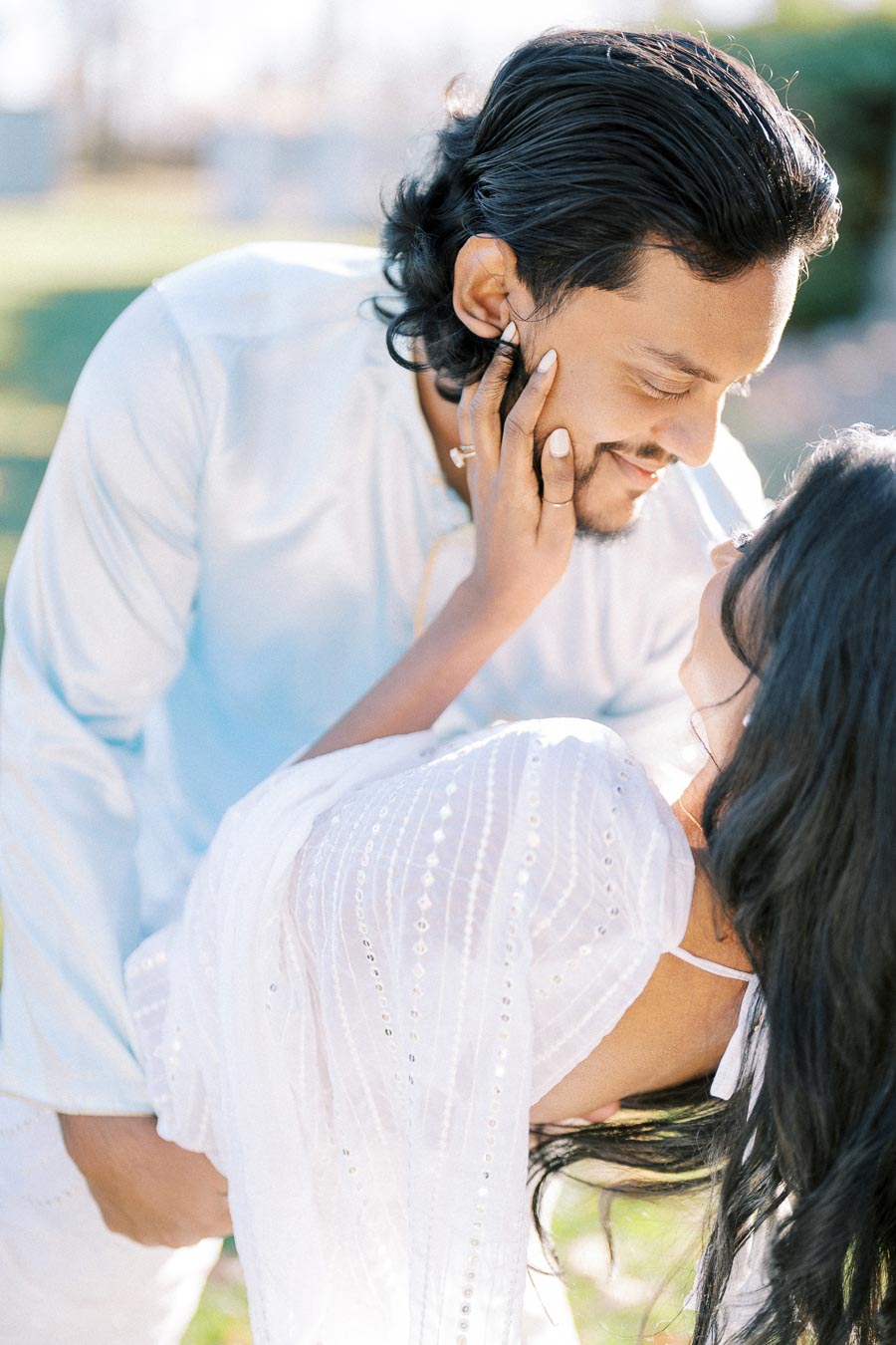 A couple sharing a romantic moment outdoors, with the woman in a white dress and the man in a light blue shirt, gazing lovingly into each other's eyes surrounded by soft, natural light.