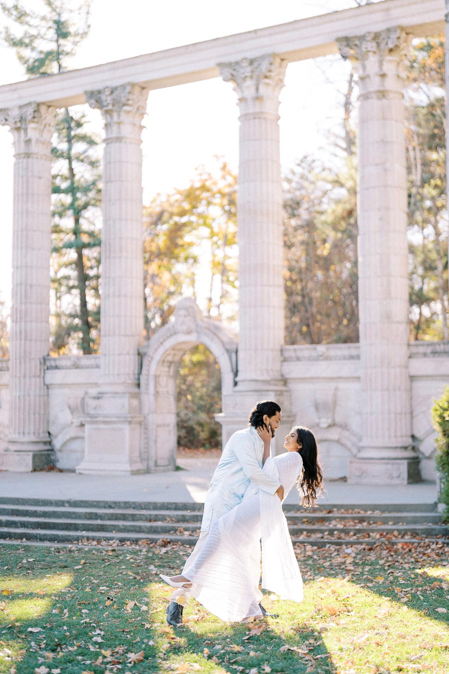 A couple in white clothing shares a romantic moment surrounded by ancient stone columns and autumn foliage, in an outdoor setting bathed in warm sunlight.