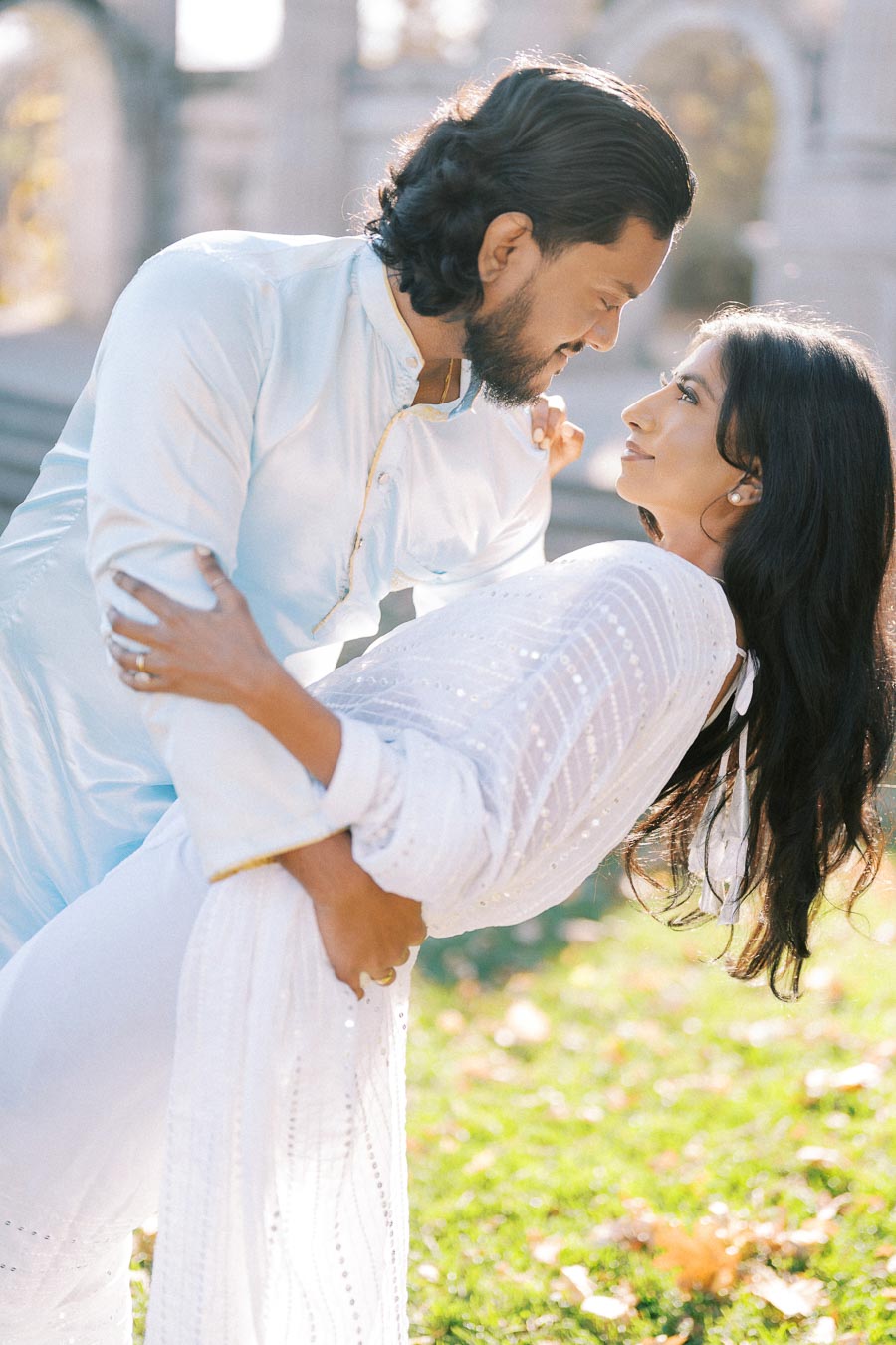 A couple in traditional white attire sharing a romantic moment outdoors, surrounded by bright sunshine and greenery, with an elegant architectural background.