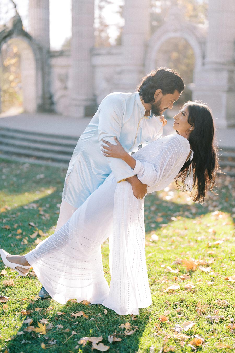 A couple dressed in elegant attire shares a romantic dance outdoors in a sunlit garden, with classical architecture in the background.