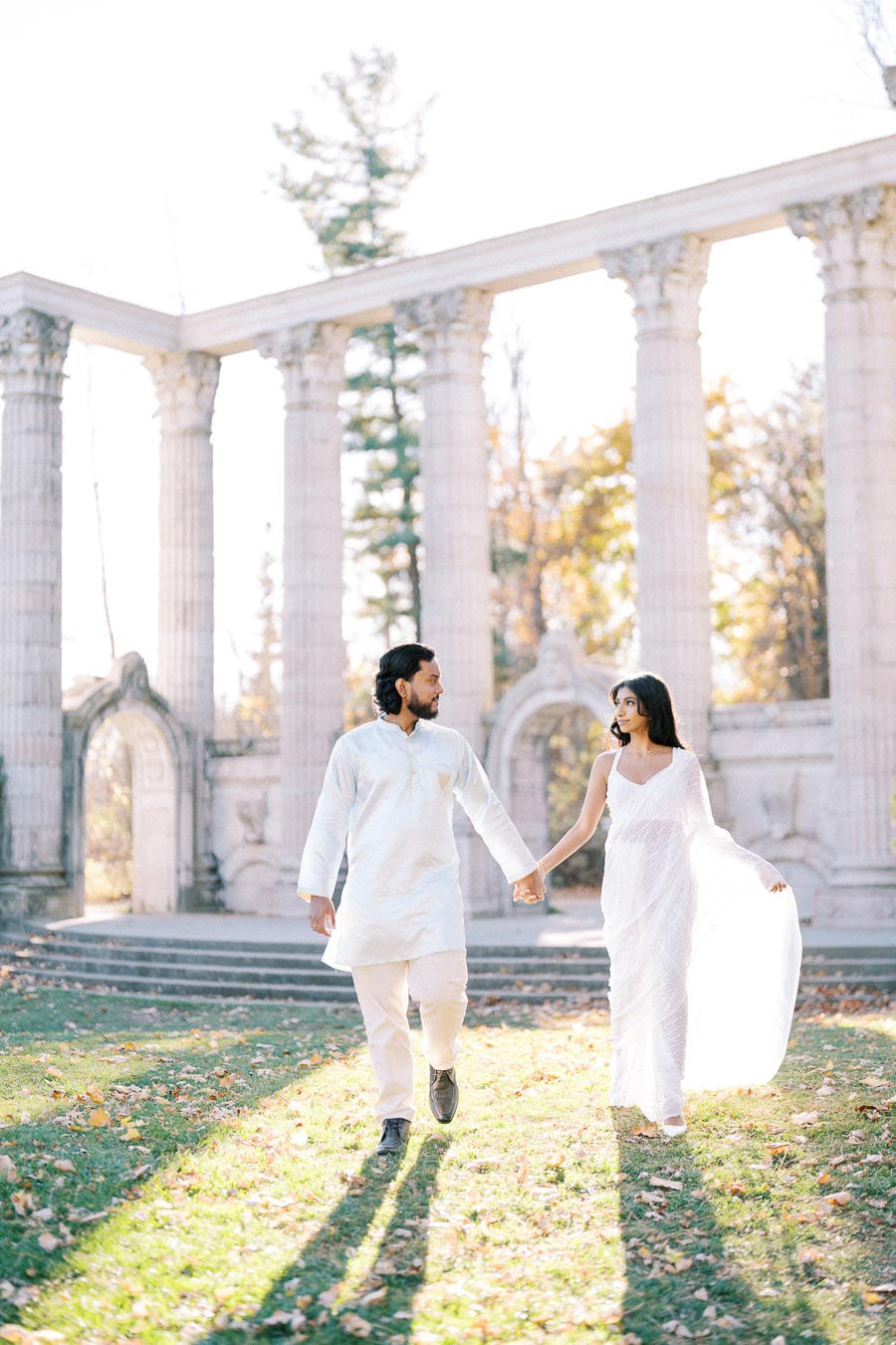 A couple in traditional white attire walking hand in hand in front of ancient stone columns with autumn foliage in the background.