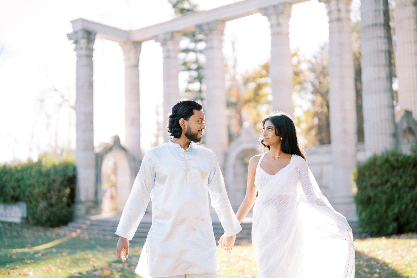A couple in elegant white attire walking hand in hand in front of ancient stone columns, with greenery and sunlight in the background.