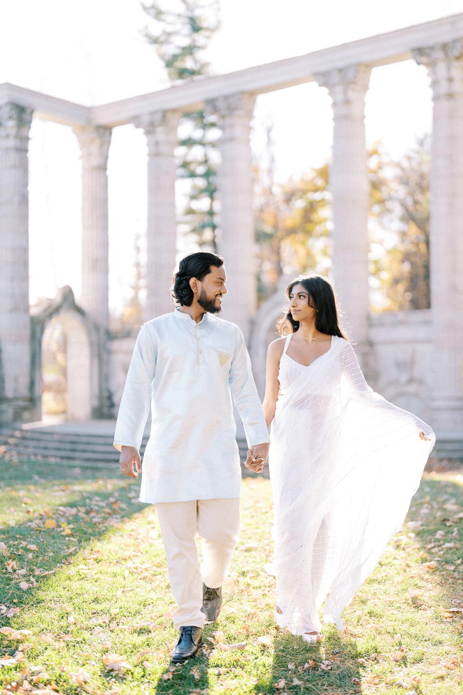 A couple in traditional white attire holding hands and walking in a sunlit garden with stone arches in the background, exuding romance and elegance.