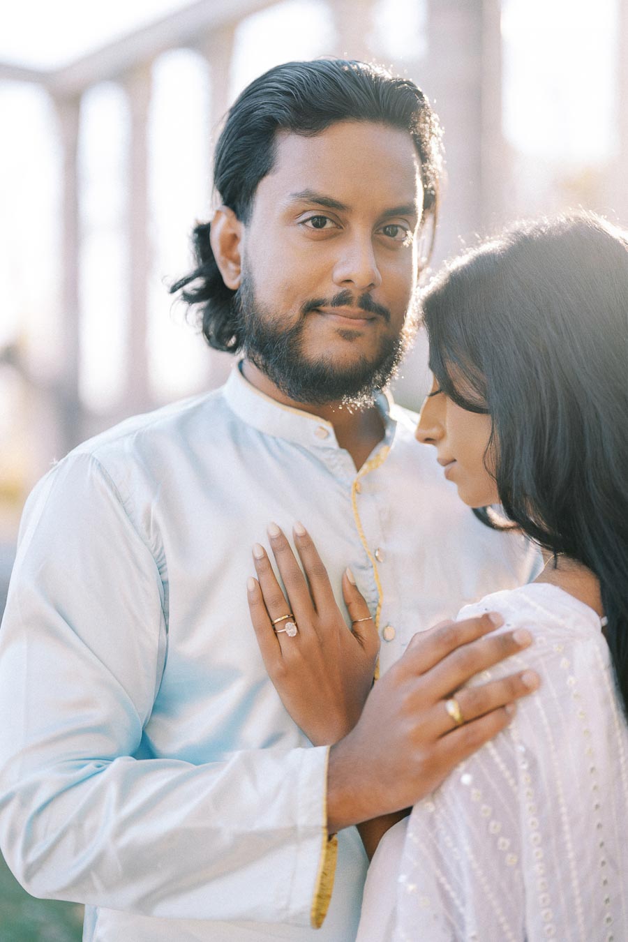 Couple embracing in soft sunlight with an elegant aqueduct in the background, showcasing engagement rings and delicate attire.