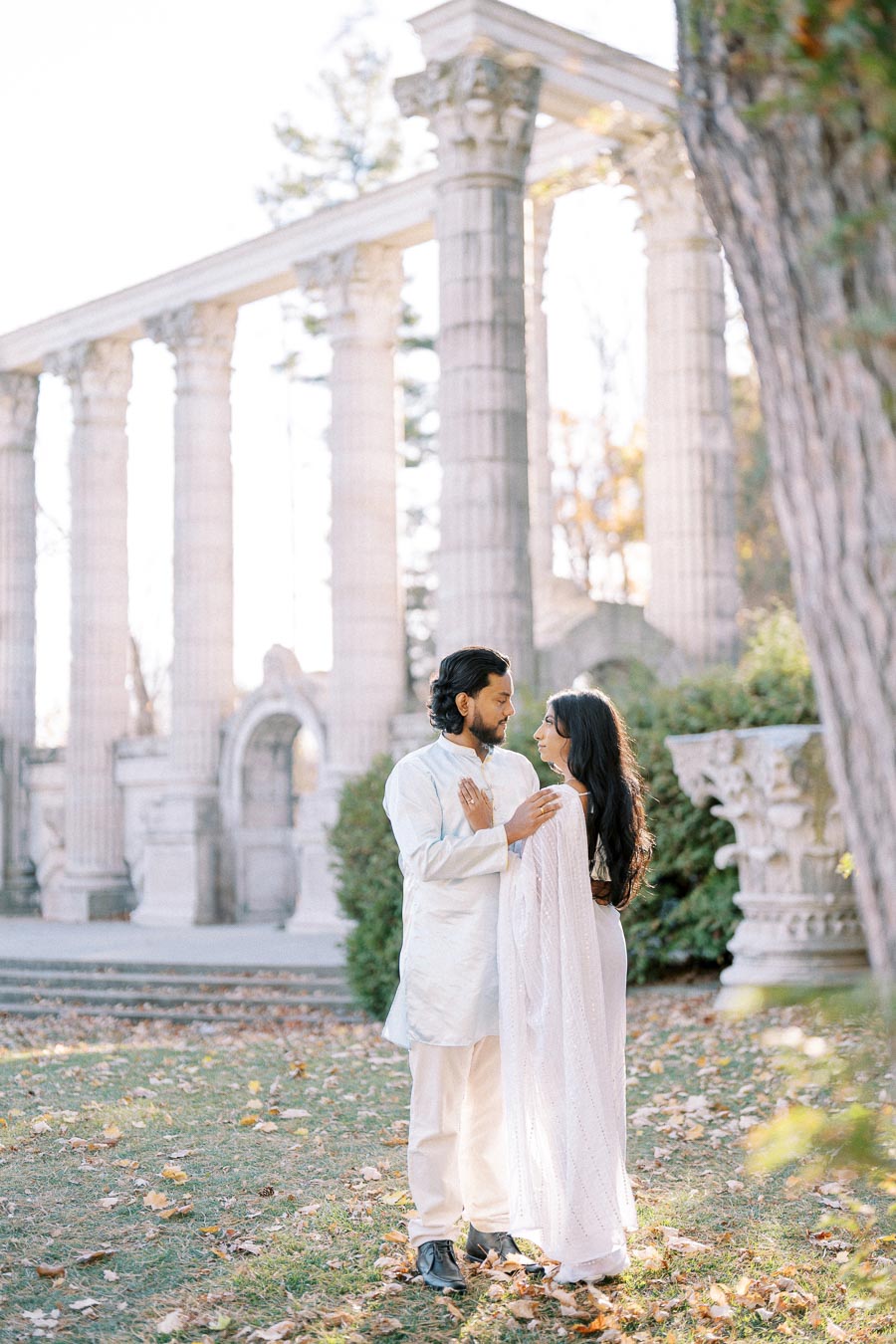 A couple embracing in front of ancient-style columns and lush greenery, wearing traditional white attire, in a serene outdoor setting.