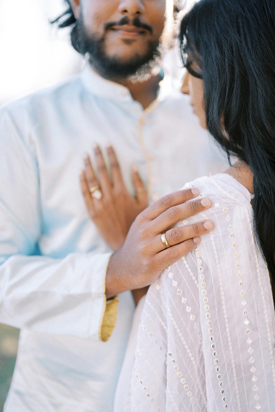 A couple standing close together, focusing on their hands with matching rings, wearing light-colored traditional clothing, symbolizing love and commitment.