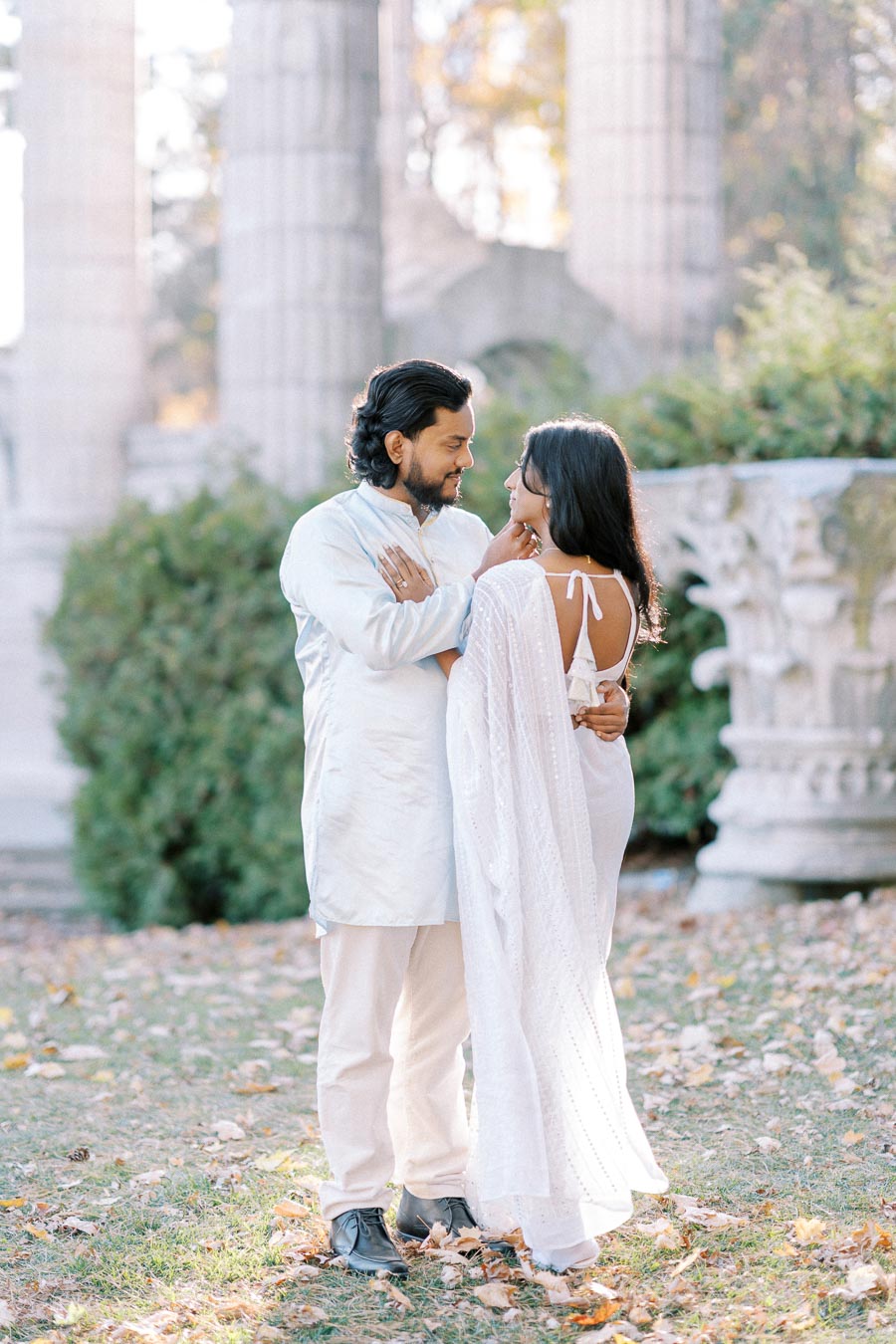 A couple in traditional white attire embraces lovingly in a serene garden setting with stone columns and autumn leaves on the ground.