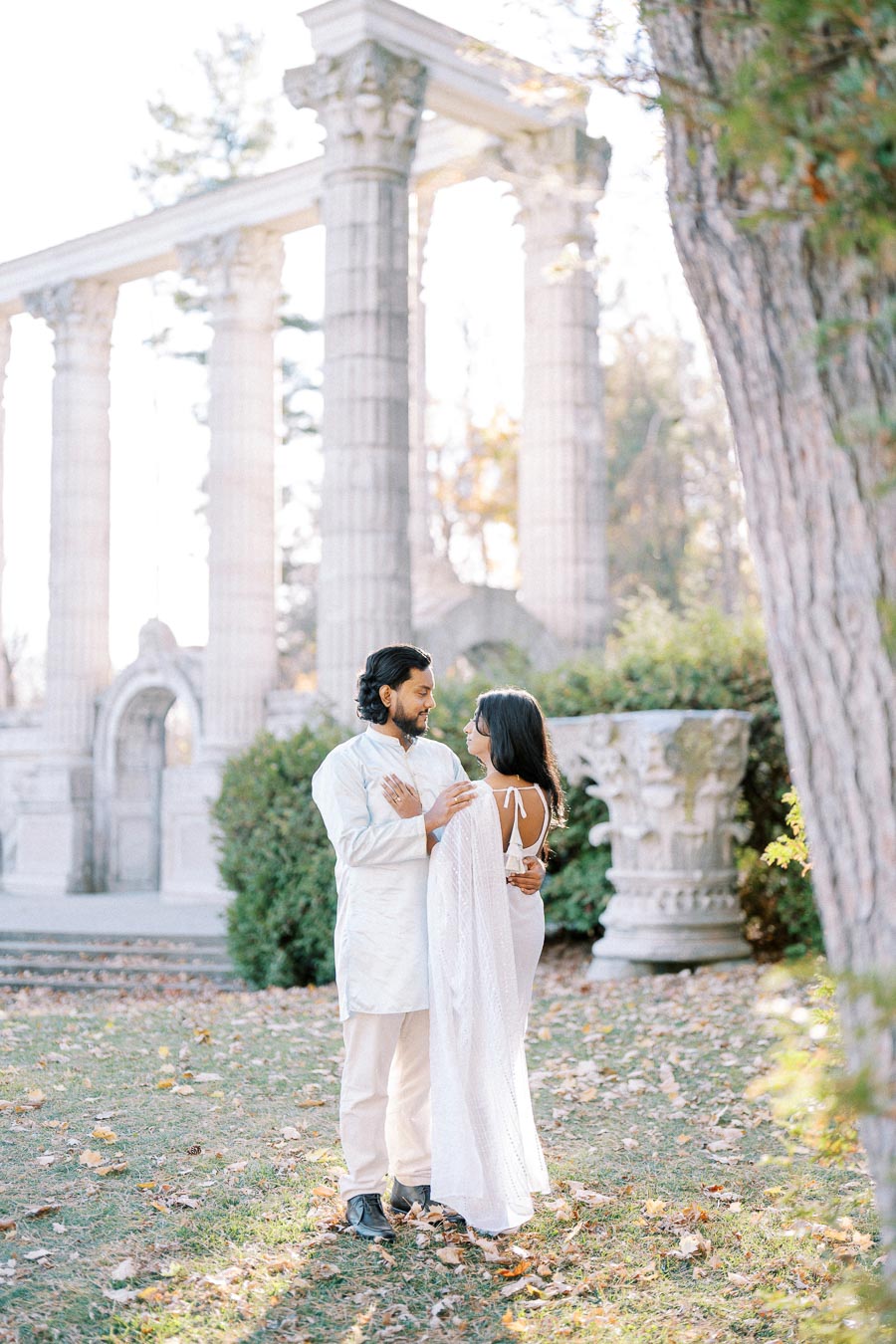 A couple in traditional Indian attire embraces in front of classical stone columns in a serene garden setting, surrounded by autumn leaves and greenery.