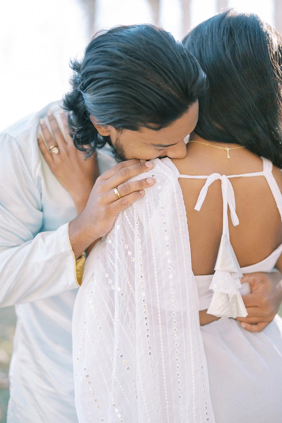 Romantic couple embracing outdoors, showcasing an intimate moment. The man gently kisses the woman's shoulder, both wearing elegant white attire, conveying affection and tenderness in a sunny setting.