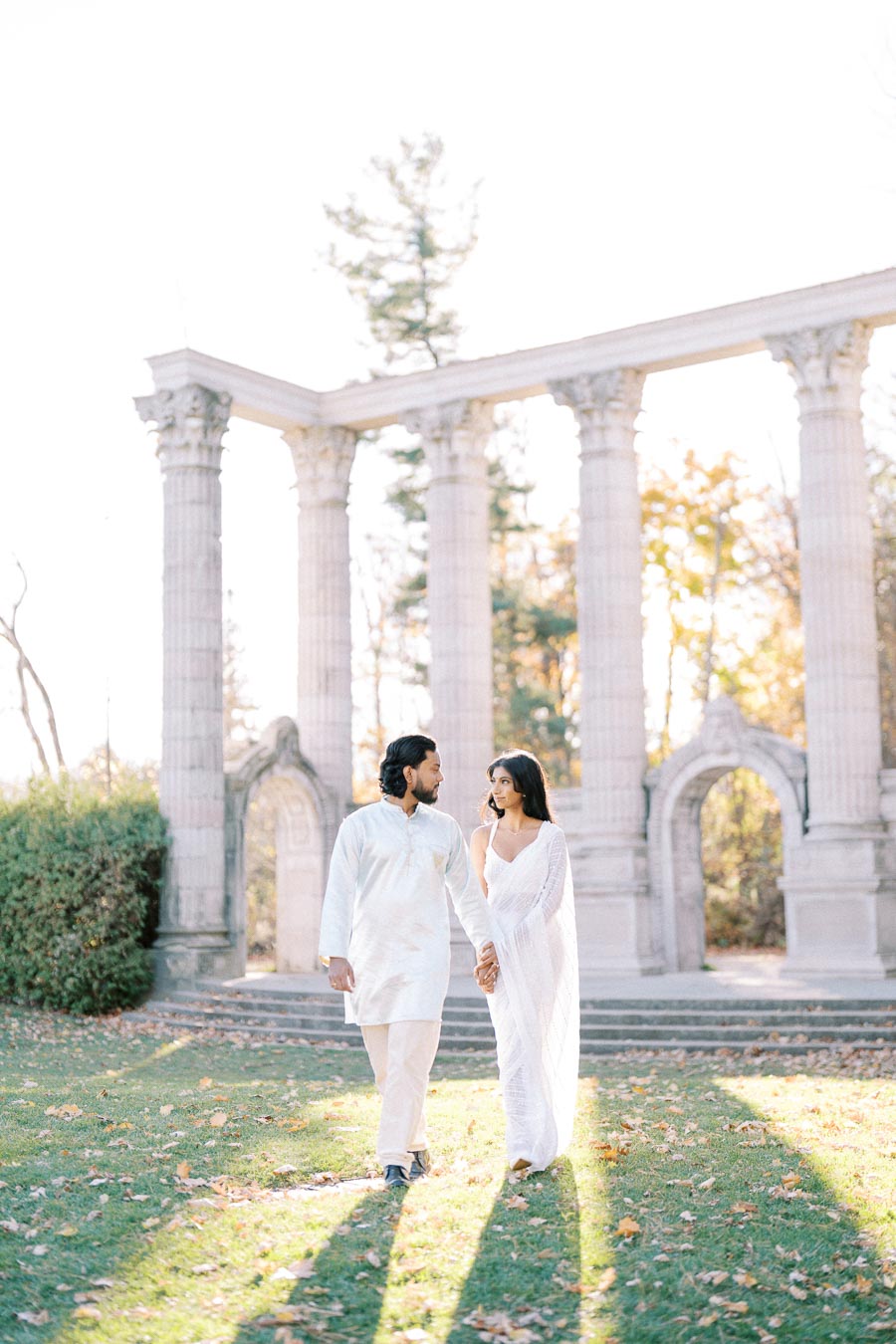 A couple in traditional attire holding hands and walking through a garden with tall stone columns in the background, surrounded by fall foliage.