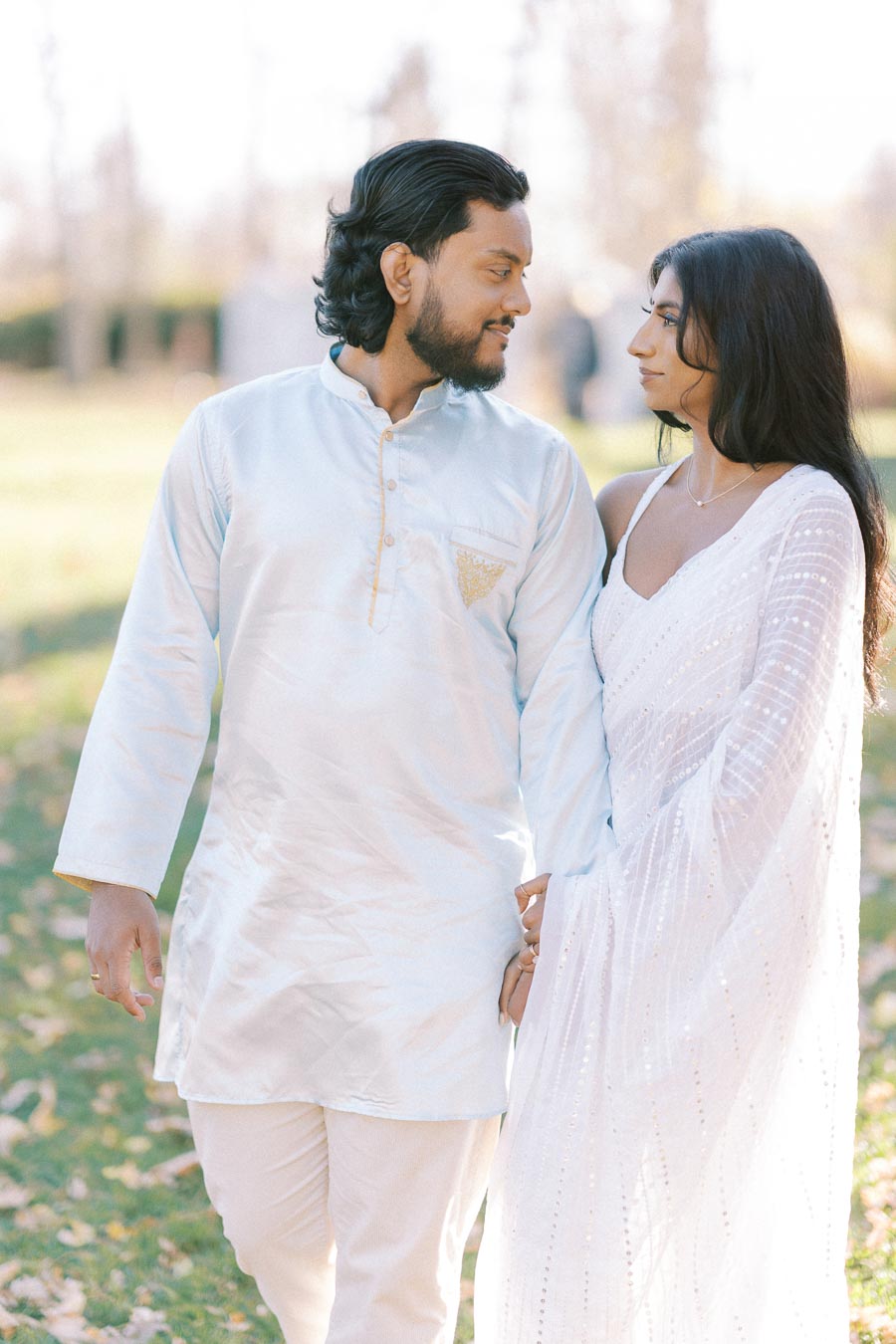A couple in traditional attire walking hand in hand on a sunny day in a park setting, with the man wearing a light blue kurta and the woman in a white saree.