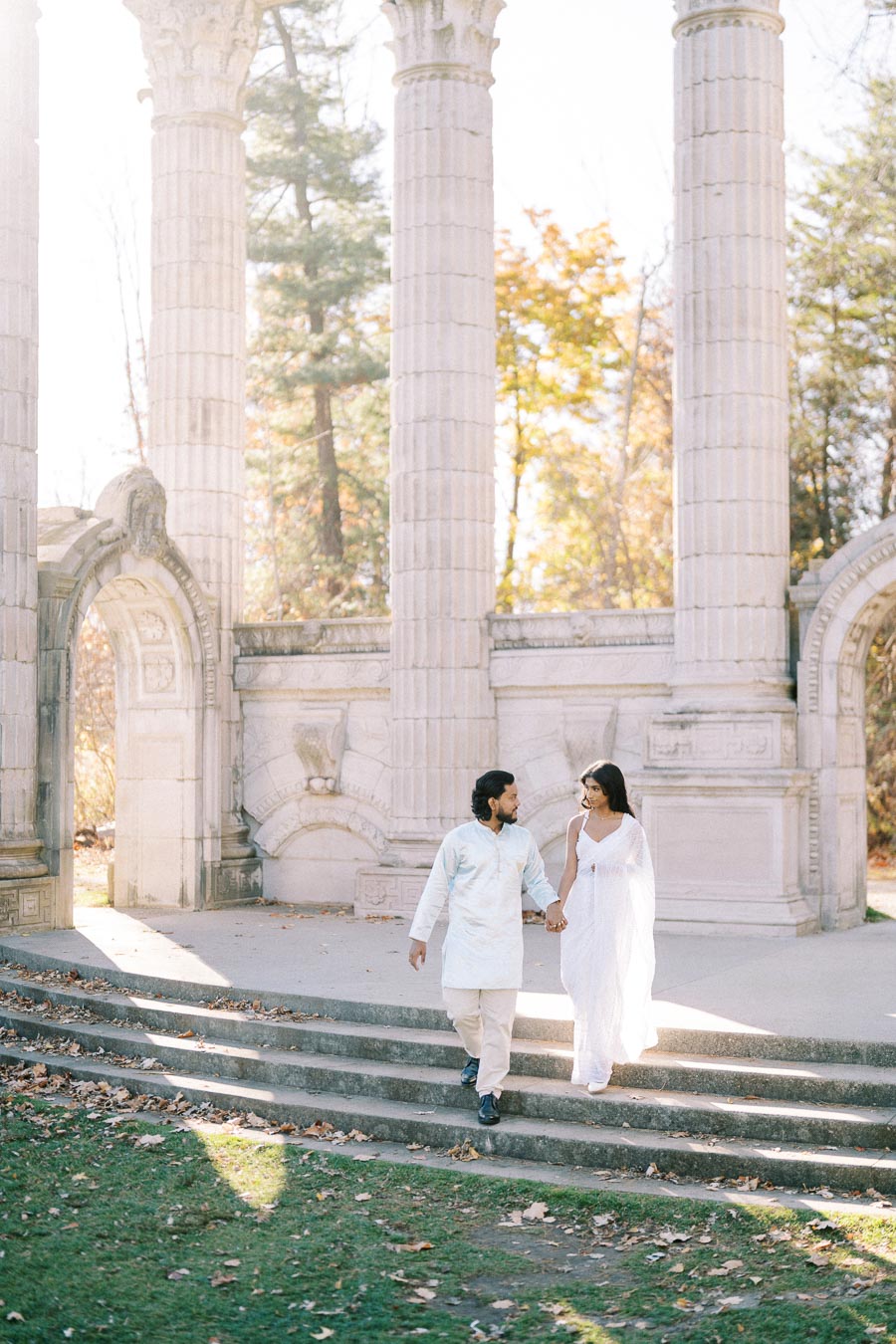 A couple wearing traditional attire walking hand in hand in front of ancient-style columns in a serene park setting.