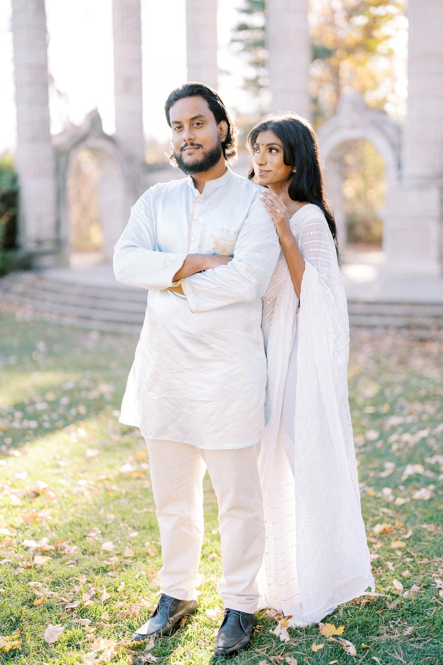 A couple dressed in traditional attire stands outside, with sunlight filtering through ancient stone columns in the background during fall.