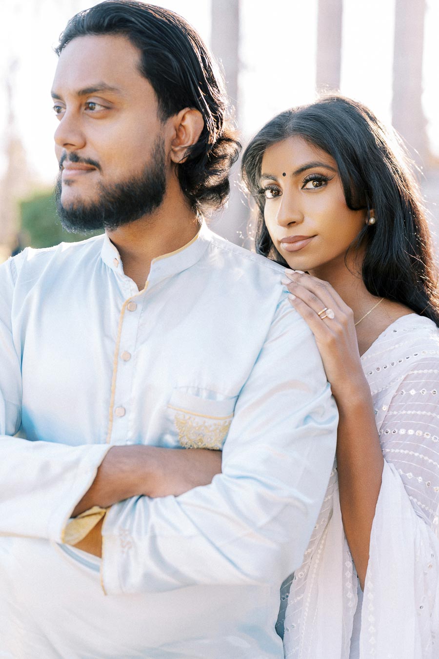 A couple posing together in traditional white attire, with the woman standing behind the man, resting her hand on his shoulder, against a soft, natural background.