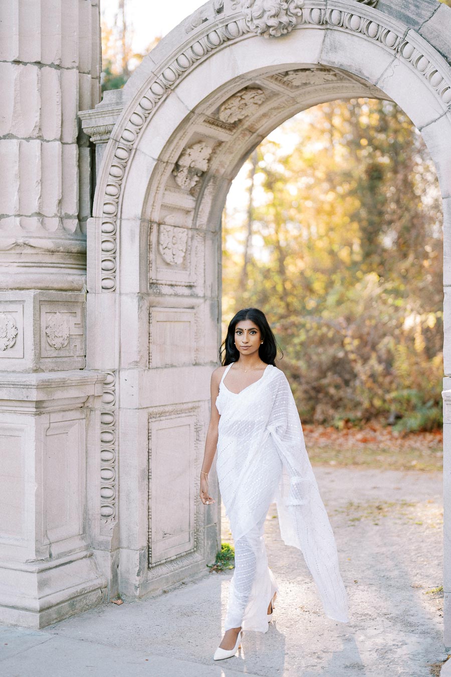 A woman in a white sari walking gracefully under an elegant stone archway, surrounded by autumn foliage.