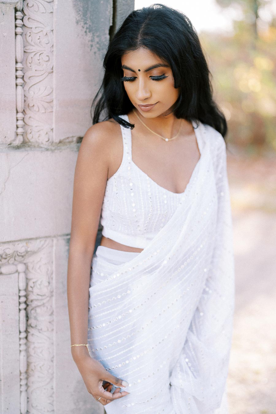 Young woman in elegant white saree standing by an intricately carved stone wall, looking downwards thoughtfully in a serene outdoor setting.