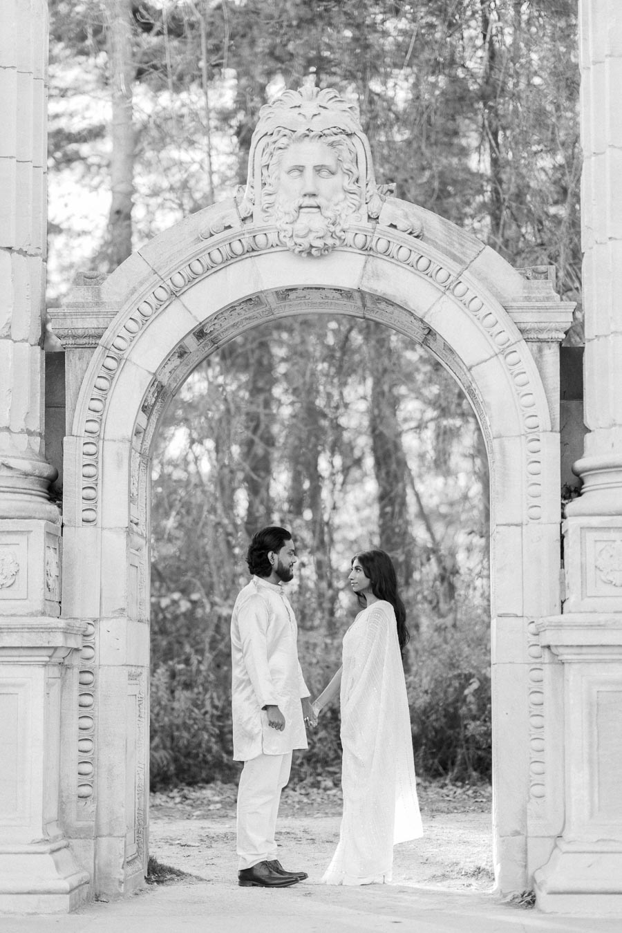 A couple stands under an ornate archway, holding hands and gazing at each other, in a serene forest setting captured in black and white.