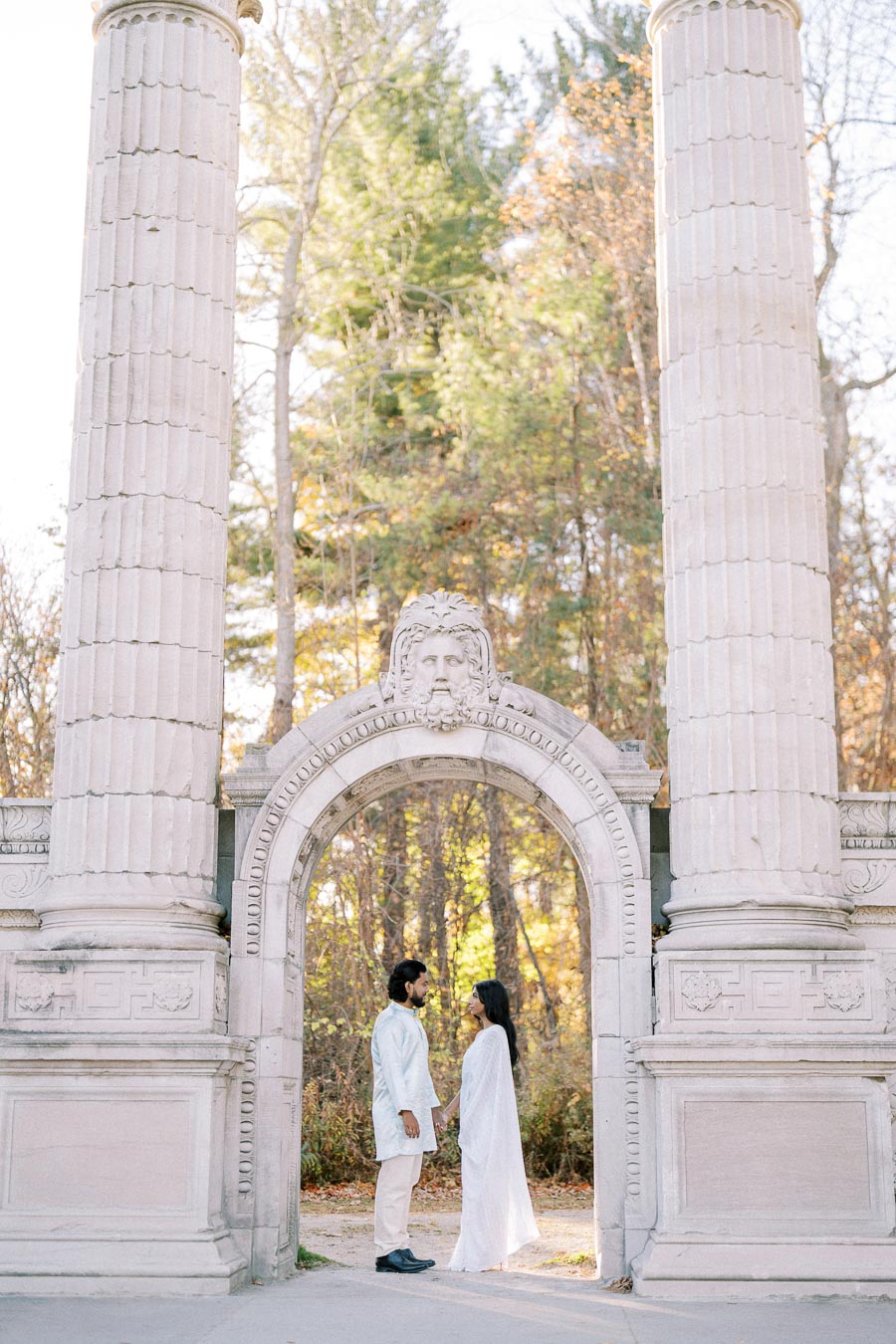 Two people stand holding hands beneath a classical stone archway with towering columns, surrounded by a lush forest in autumn.