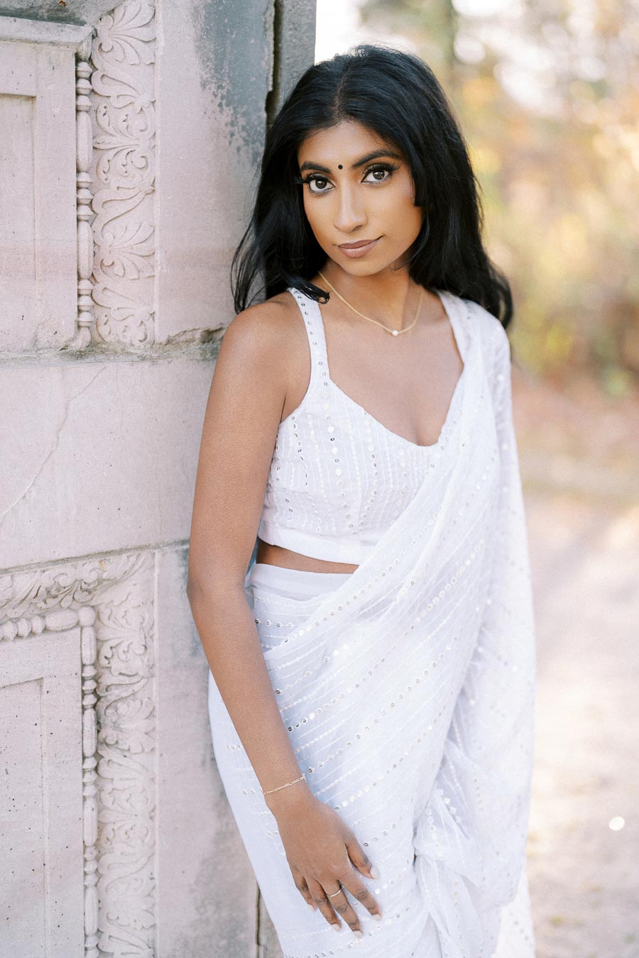 Young woman in a white sari leaning against an intricately carved stone wall, outdoors with a blurred natural background.