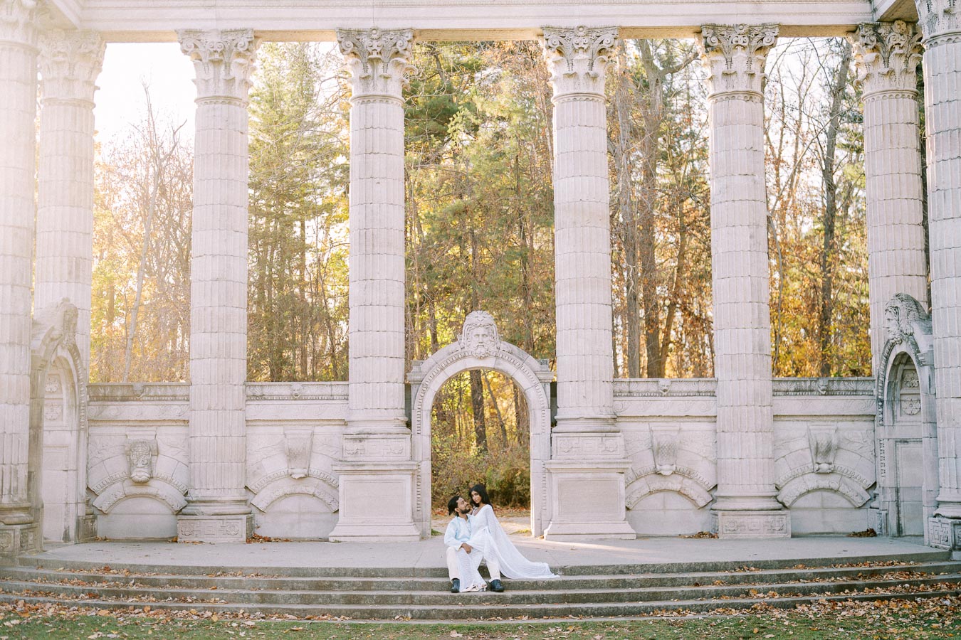 A couple in elegant white attire sits on stone steps in front of grand classical columns, surrounded by a serene autumn forest backdrop.
