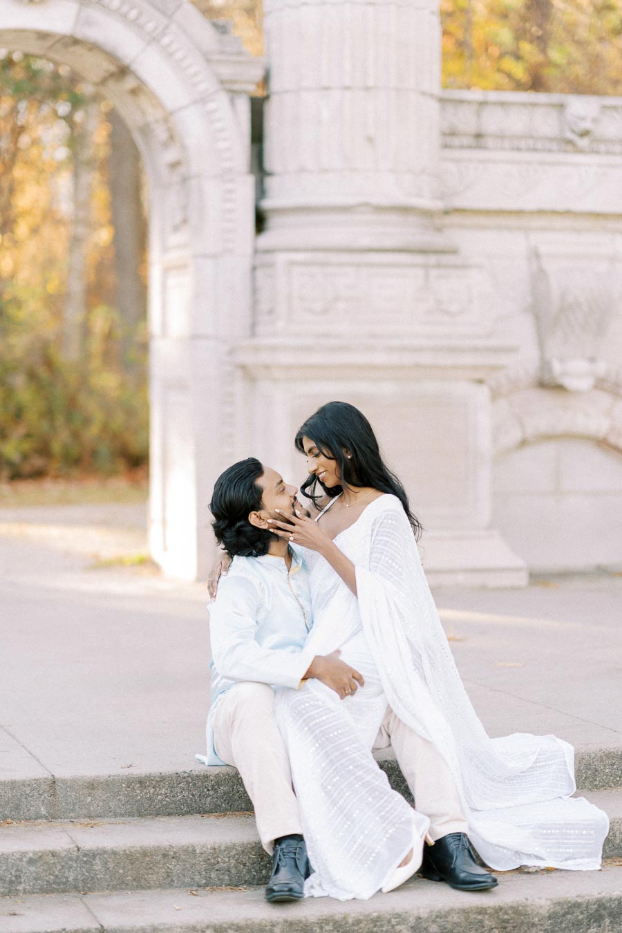 A romantic couple wearing white and light-colored attire sit on stone steps in an outdoor setting with historic architecture, softly lit by the warm glow of a sunny day, surrounded by the fall colors of the trees.