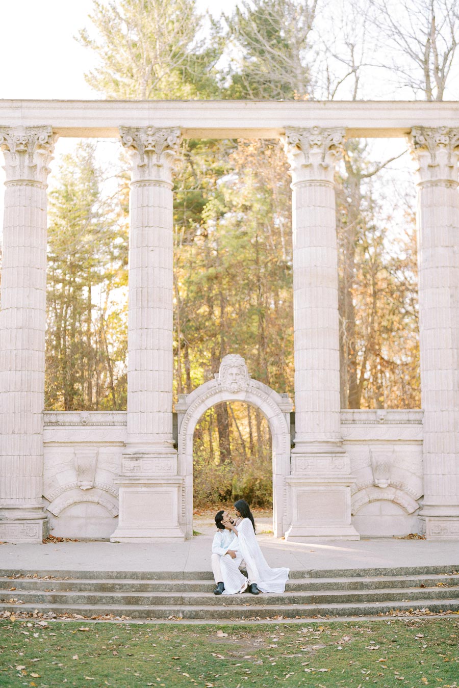 A couple in white outfits sitting on stone steps in front of ancient-style pillars and arch, surrounded by autumn trees.