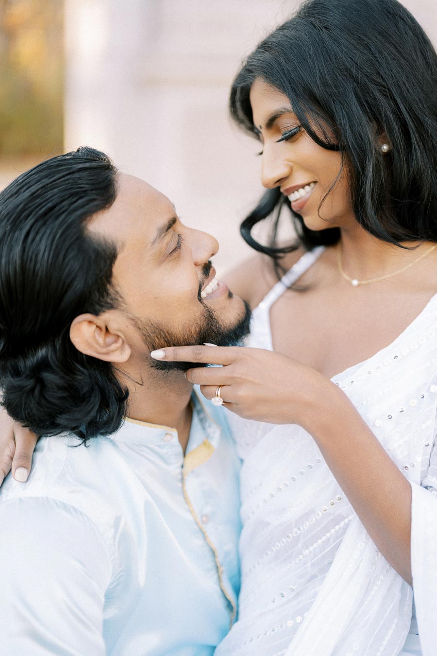 A couple gazing lovingly at each other, the woman in a white saree with intricate details, showcasing an engagement ring, as they share an intimate moment in a bright, outdoor setting.