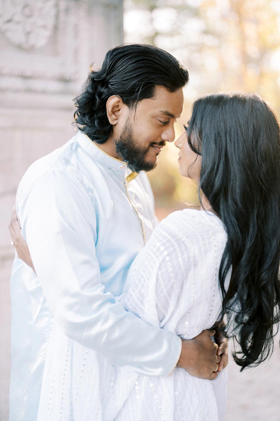 A couple dressed in elegant traditional attire embraces lovingly, exchanging a tender gaze against a soft, blurred background of outdoor scenery.