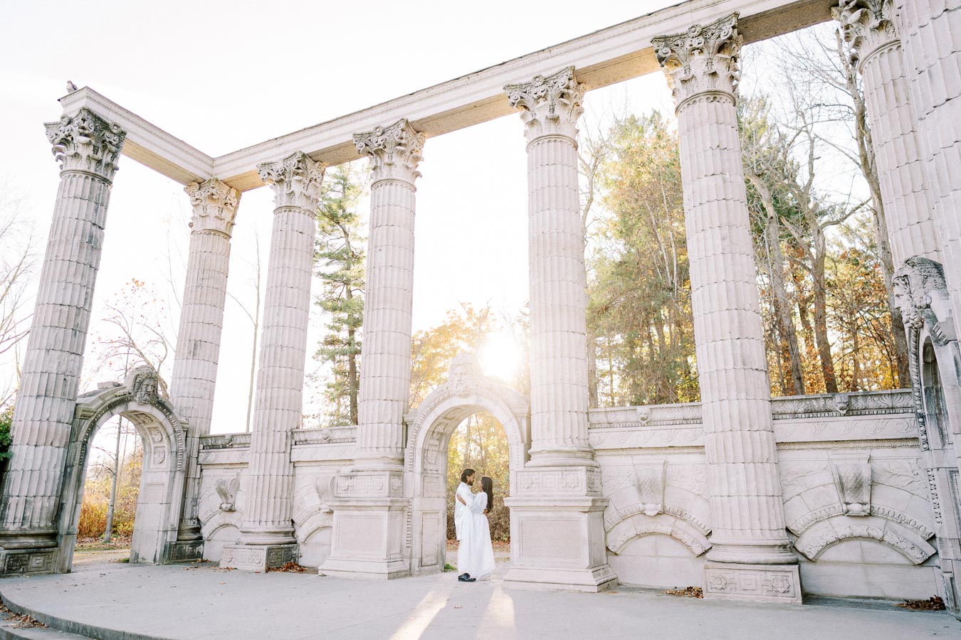 Couple embracing under ancient stone columns with sunlight filtering through, surrounded by autumn trees at a historic architectural site.