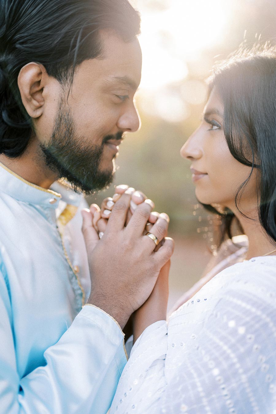 A romantic couple holding hands and gazing at each other lovingly, illuminated by soft sunlight in a serene outdoor setting.