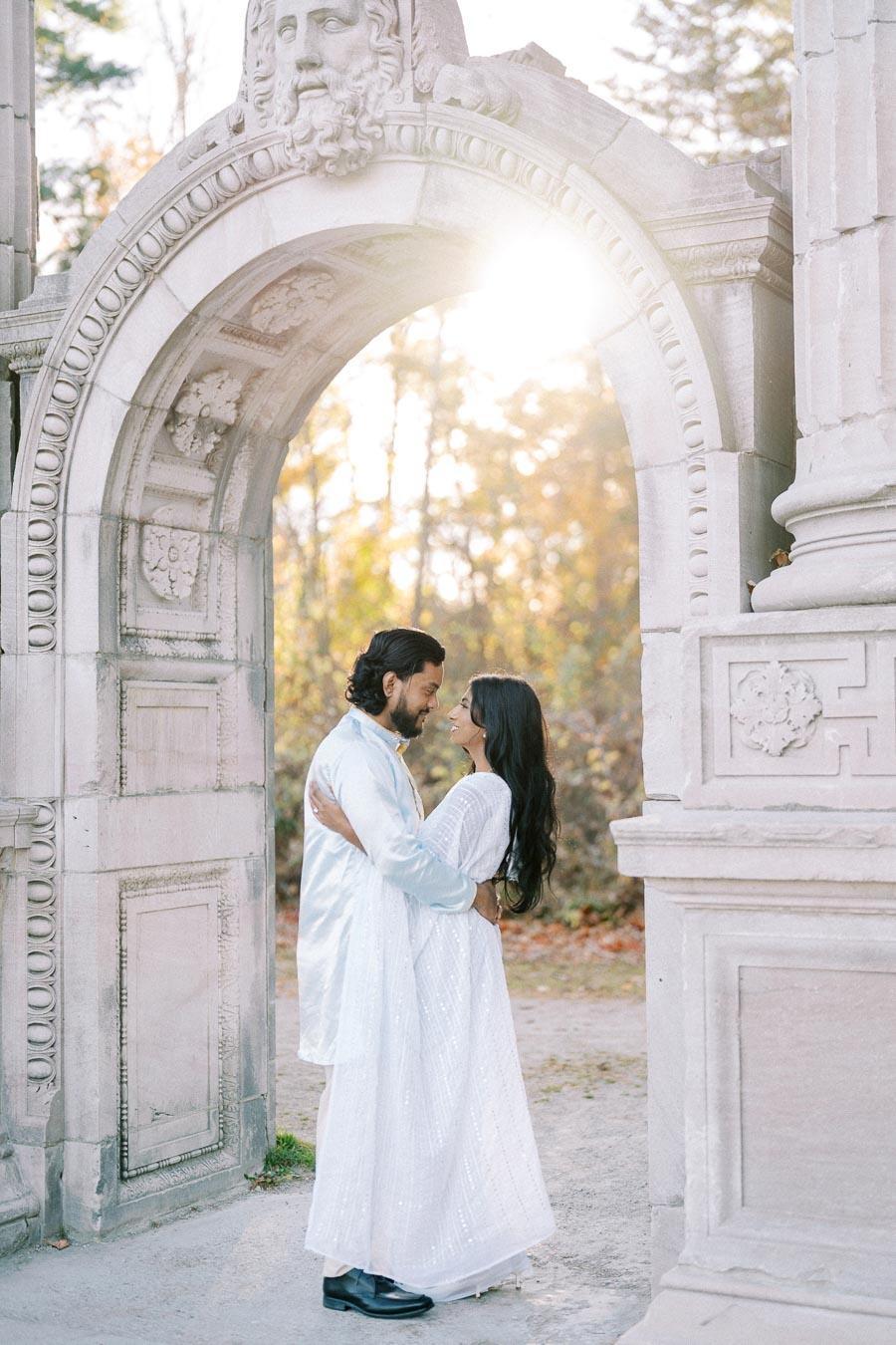 A couple embraces under a historic stone archway with sunlight filtering through, surrounded by autumn foliage.