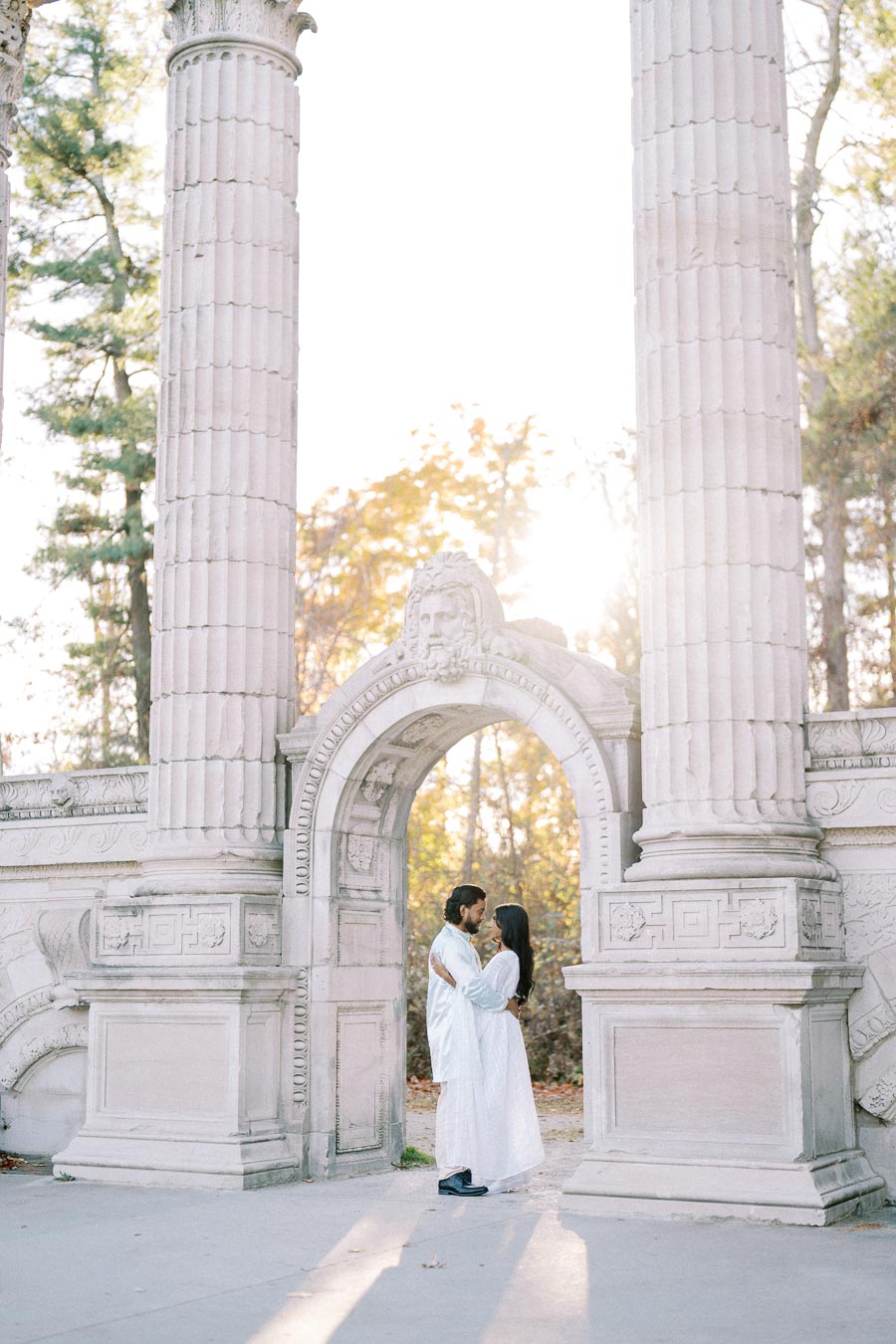 A couple embraces under a historic stone archway with ornate columns, surrounded by autumn foliage, as sunlight filters through the trees.