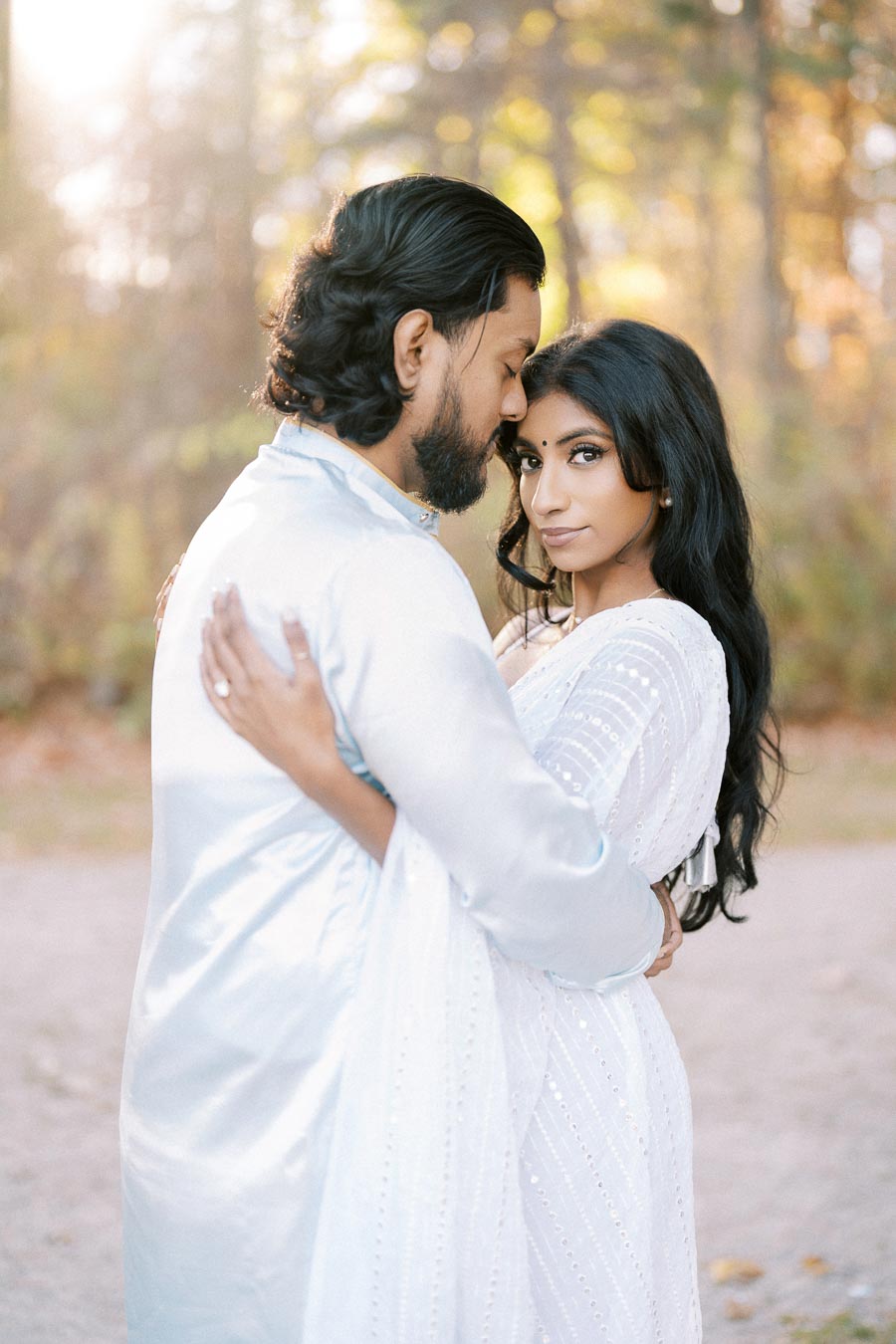A couple embracing in a serene outdoor setting, dressed in matching white traditional attire, with soft sunlight filtering through the trees in the background.