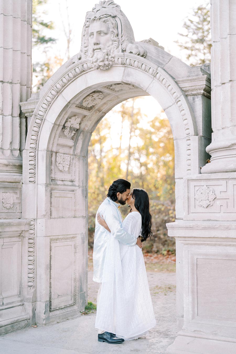 A couple dressed in white embraces under an ornate stone archway with a carved face at the top, surrounded by autumn trees.
