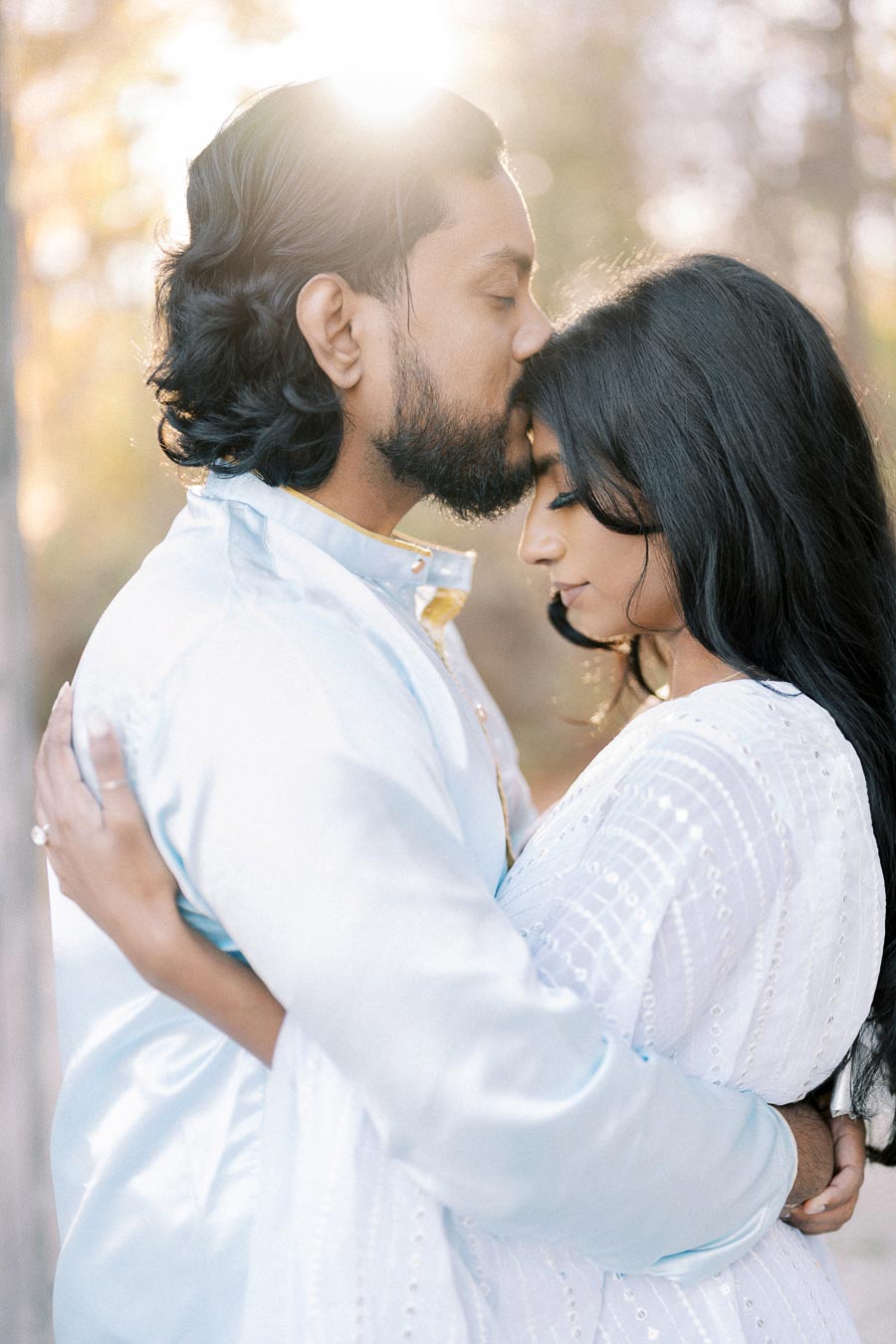 A couple embraces romantically in a sunlit forest, both wearing soft blue attire.