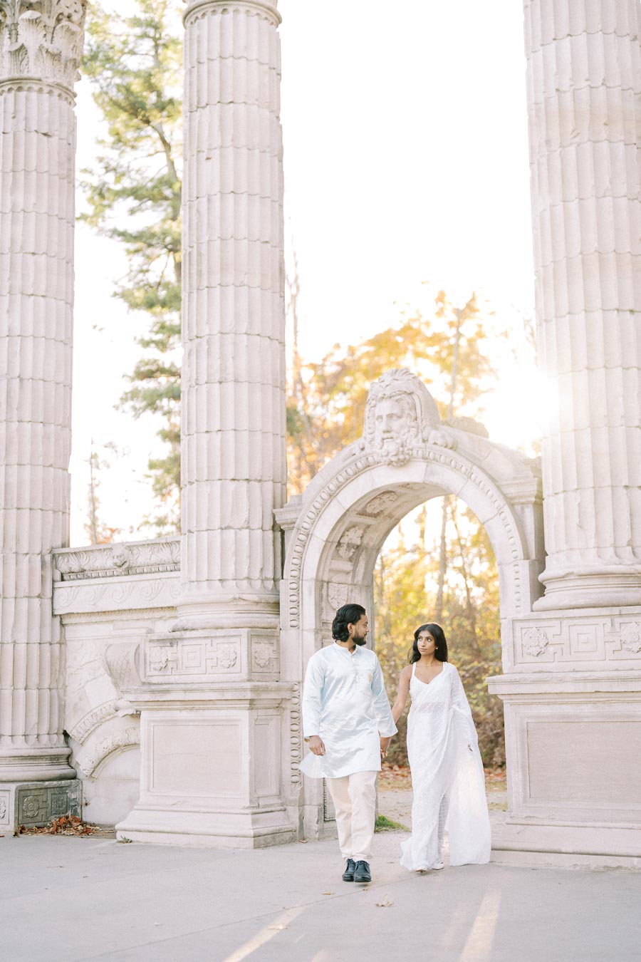 Couple walking hand in hand through historic stone archway with autumn foliage.