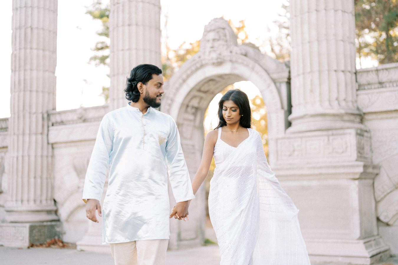 A couple dressed in traditional South Asian attire holding hands while walking near an ancient stone archway with intricate columns.