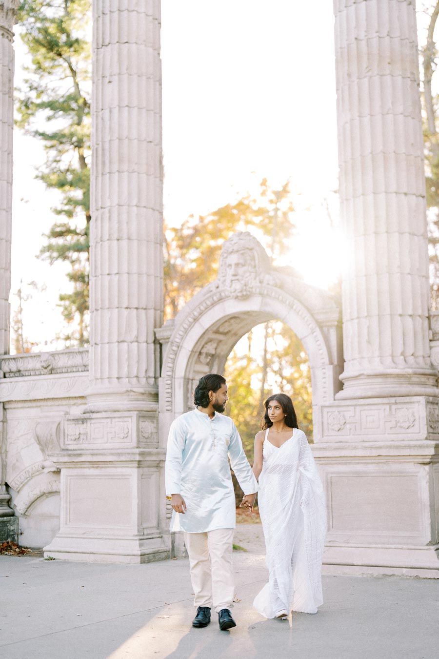 A couple holding hands and walking in front of historic stone columns and archway, with sunlight streaming through trees in the background.