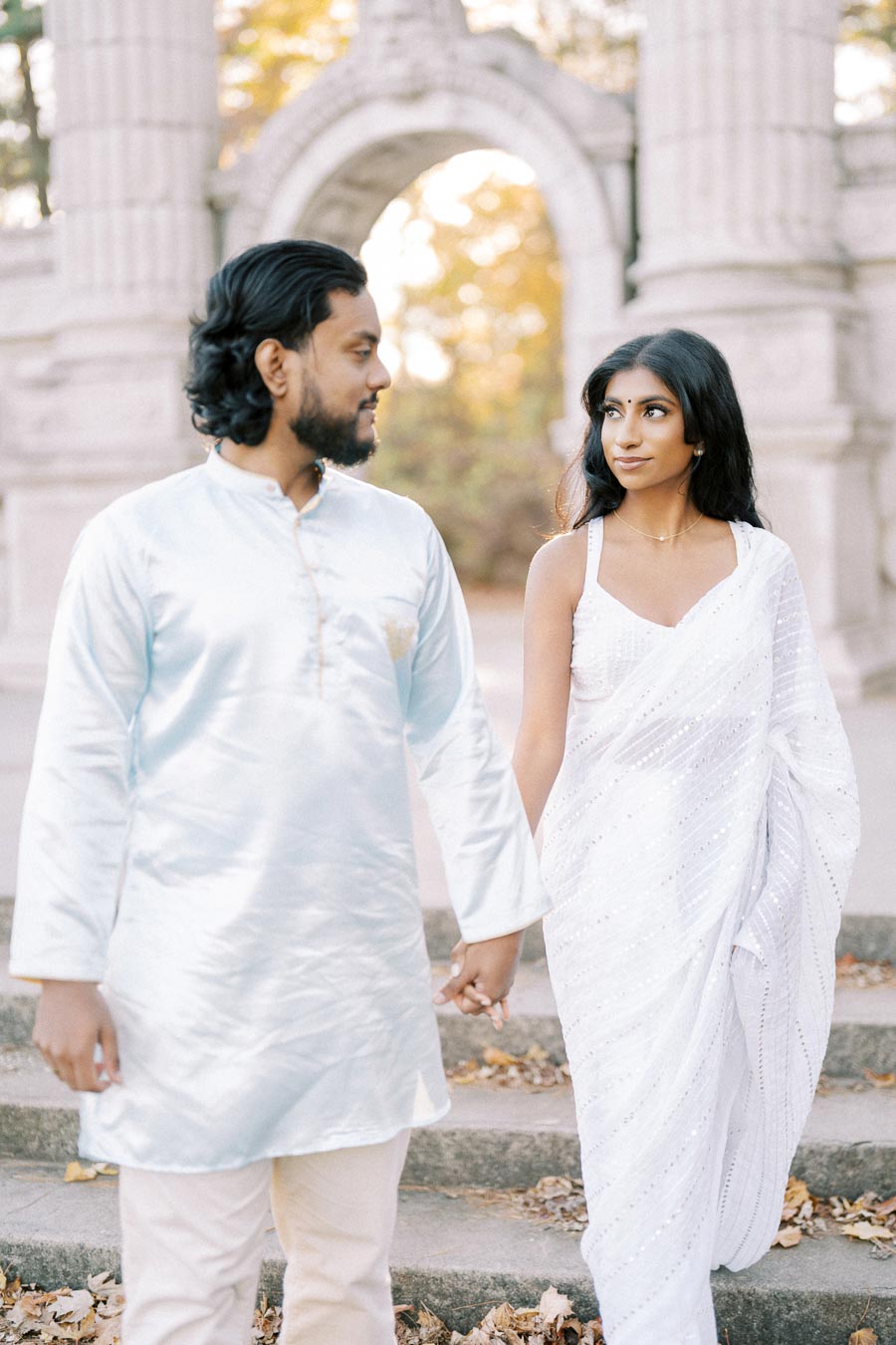 A couple dressed in elegant white traditional attire holding hands and walking on stone steps with an ancient stone arch in the background.