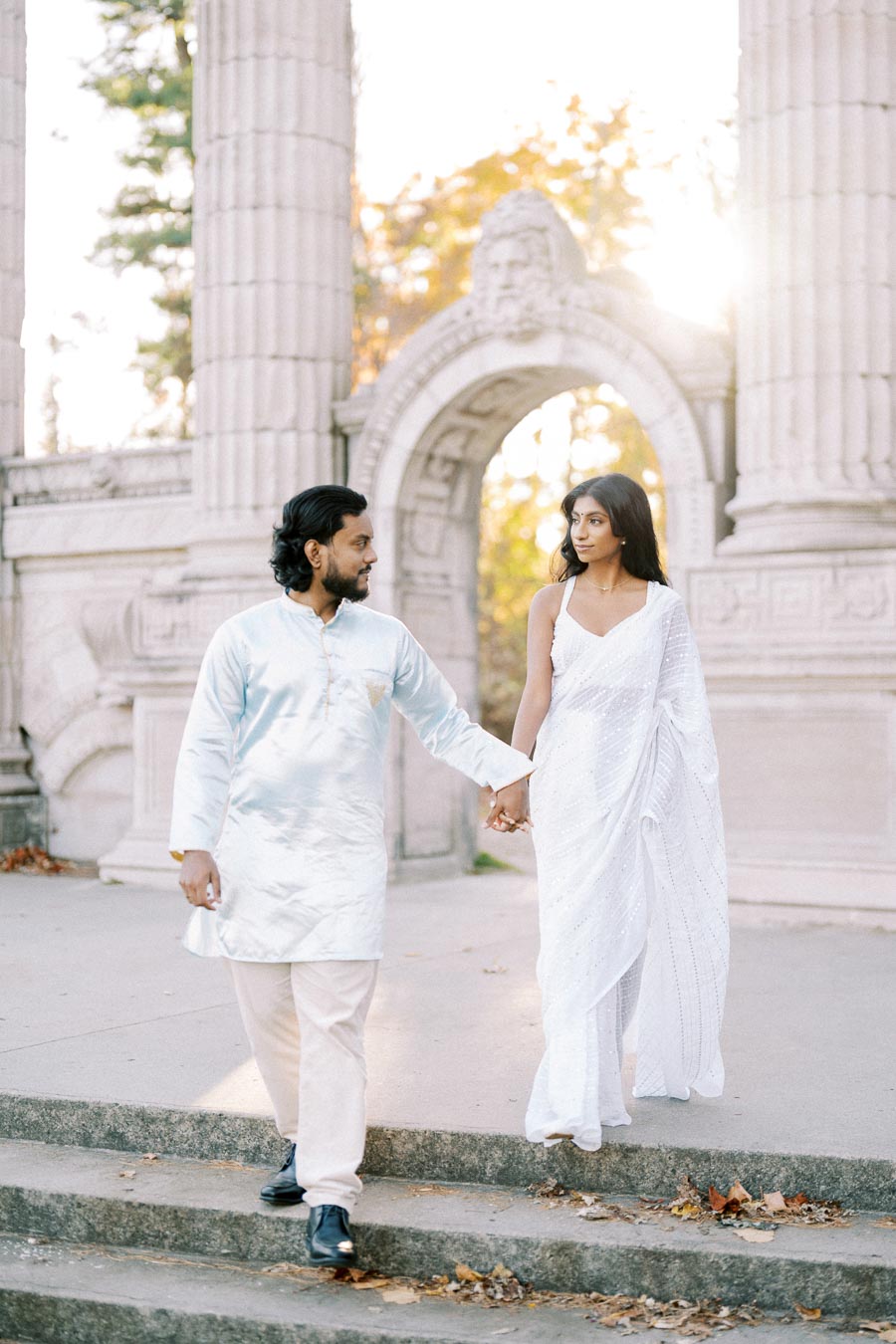 A couple in traditional attire holding hands and walking down steps in front of historic stone arches, with sunlight filtering through the trees in the background.