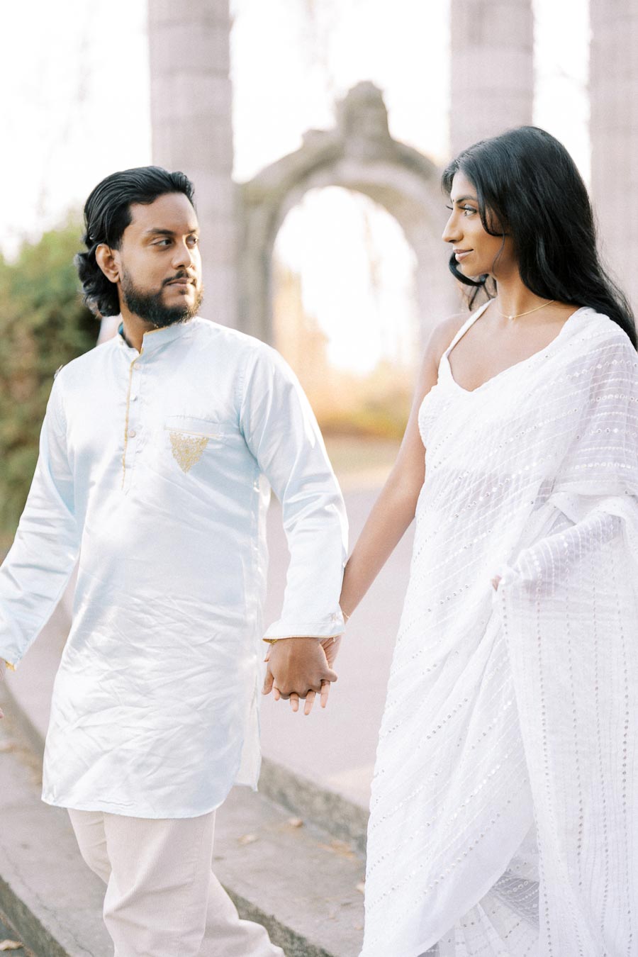 A couple in traditional attire holding hands and walking outdoors, with an ancient architectural backdrop, symbolizing love and unity in a cultural setting.