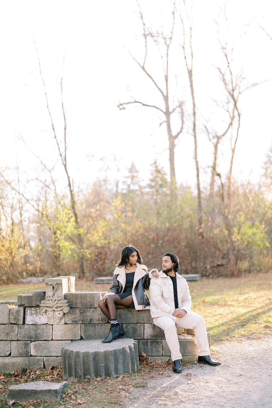 A couple sitting on stone blocks in a park during autumn, surrounded by bare trees and sunlight, wearing stylish fall outfits.