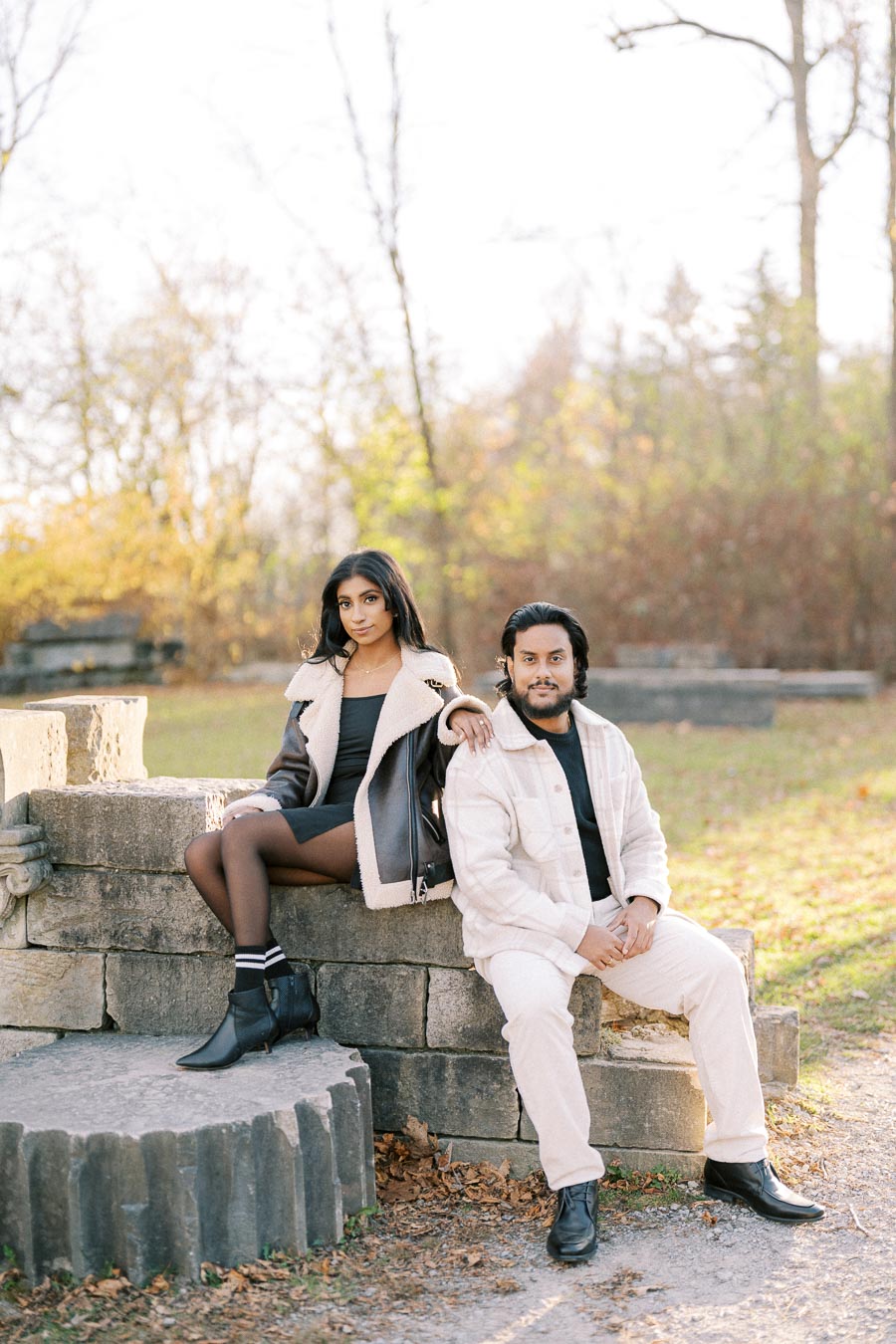 A couple posing outdoors on a stone bench in autumn, with the woman wearing a shearling jacket and black boots and the man in a light jacket and black shoes. Trees with fall foliage can be seen in the background.